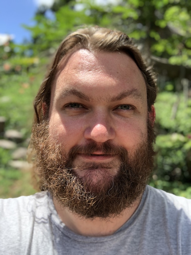 Picture of man with beard and long hair wearing grey shirt, smiling in front of tropical foliage